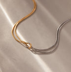 The Mixed Metal Knot Necklace features intertwined gold and silver chains, elegantly displayed on a soft beige surface under gentle lighting—a refined symbol of everlasting love.