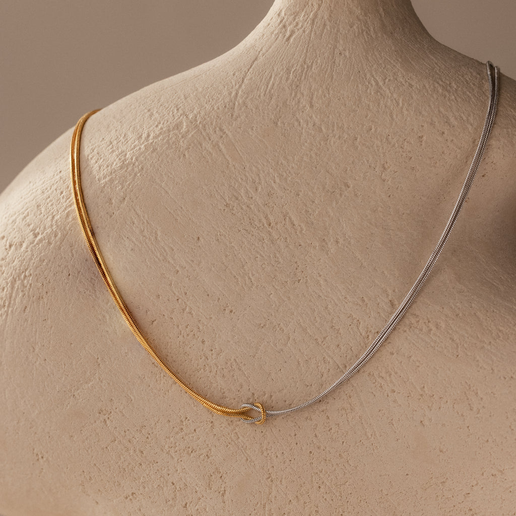 Two-tone gold and silver chains grace a textured mannequin, highlighting the Mixed Metal Knot Necklace—a stunning symbol of everlasting love.