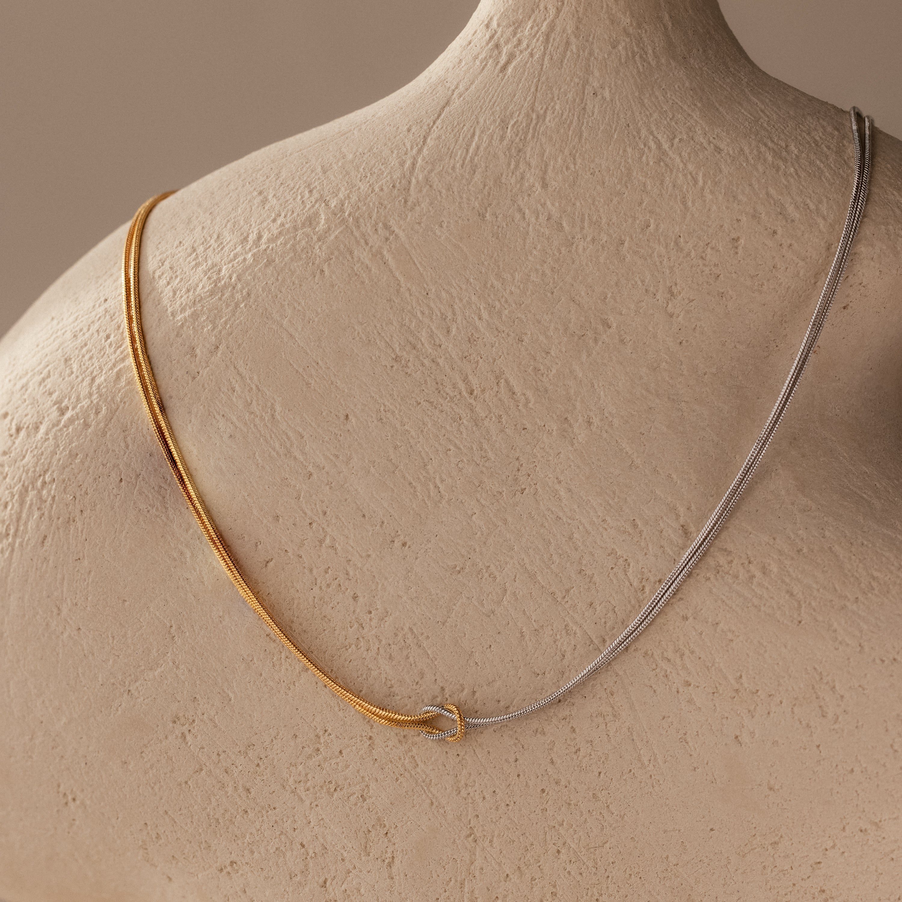 Two-tone gold and silver chains grace a textured mannequin, highlighting the Mixed Metal Knot Necklace—a stunning symbol of everlasting love.