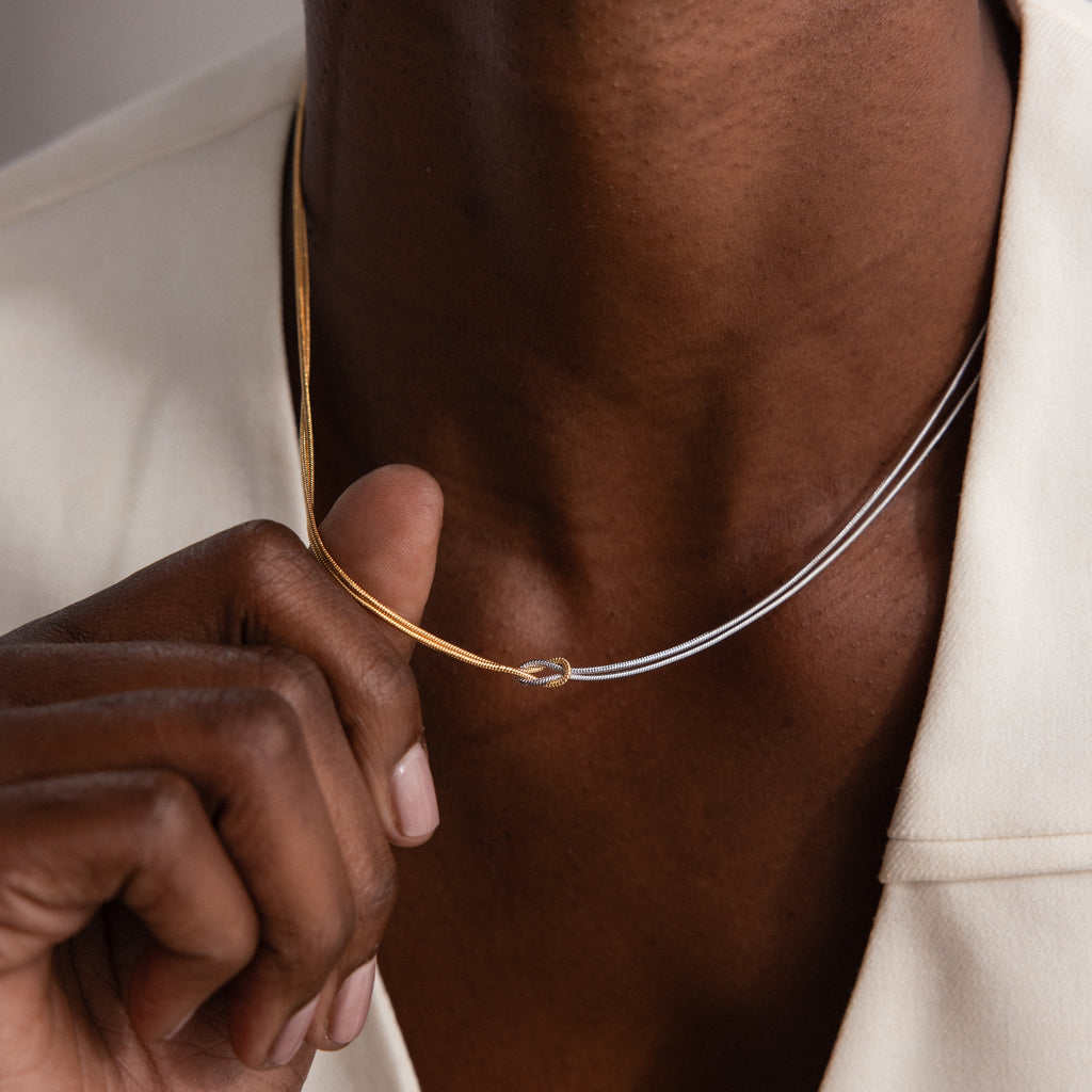 A person in a cream blazer holds the Love Knot Chain Necklace, featuring gold and silver tones, close to their neck.