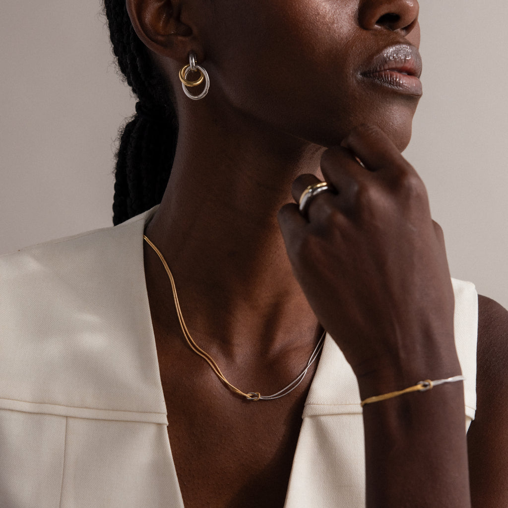 Wearing the Mixed Metal Knot Necklace along with gold and silver earrings, bracelet, and ring, a woman in a cream sleeveless top radiates everlasting love.