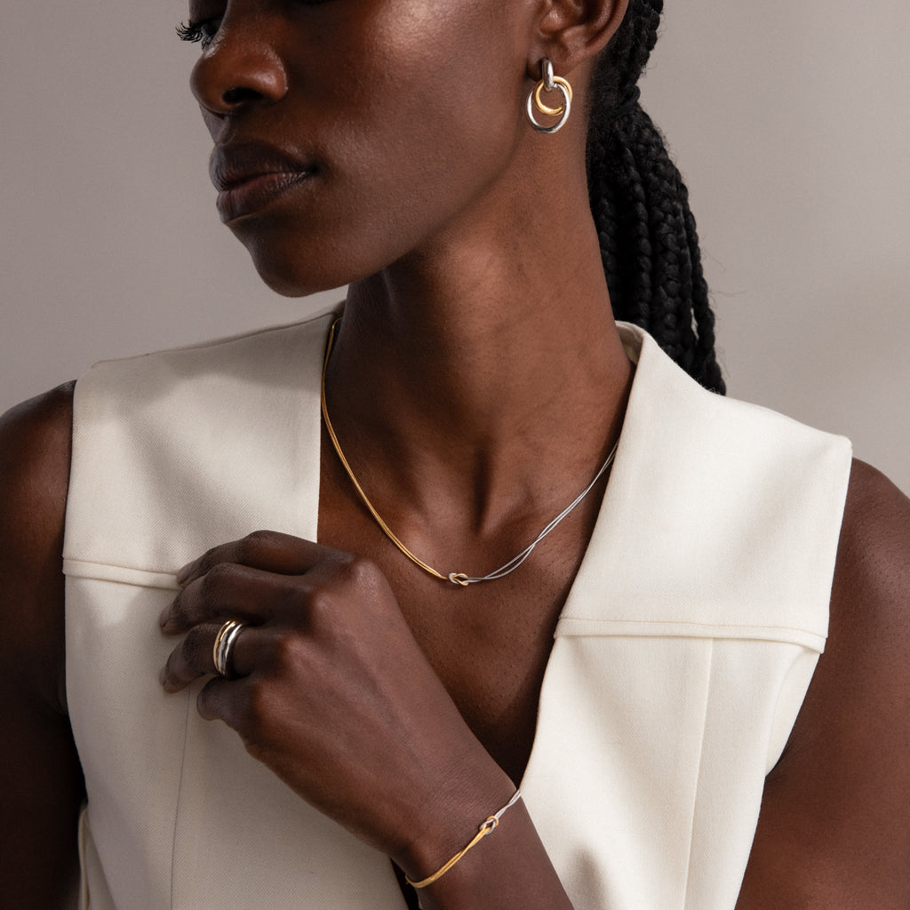 A woman poses with her hand near her collarbone, wearing a gold and silver Love Knot Chain Necklace over a white sleeveless top.
