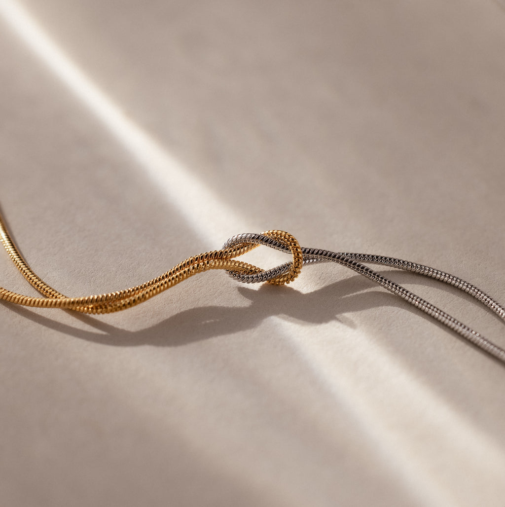 The Mixed Metal Knot Necklace features intertwined gold and silver chains knotted together, beautifully symbolizing everlasting love on a softly lit, neutral background.