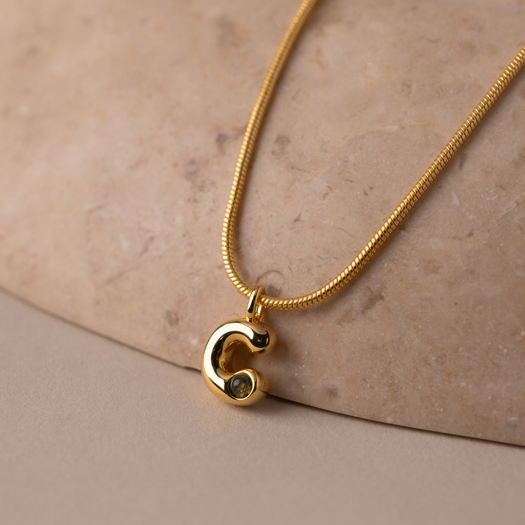 Gemstone Bubble Initial Necklace with a C-shaped pendant, elegantly displayed on a smooth beige stone—a timeless personalized jewelry piece.