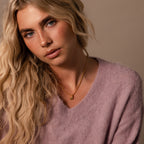 A woman with long blonde hair, wearing a soft pink sweater and the Gemstone Bubble Initial Necklace, looks at the camera.