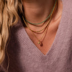 Close-up of a woman wearing a mauve sweater and layered gold and green bead necklaces, featuring the Gemstone Bubble Initial Necklace for a personalized touch.