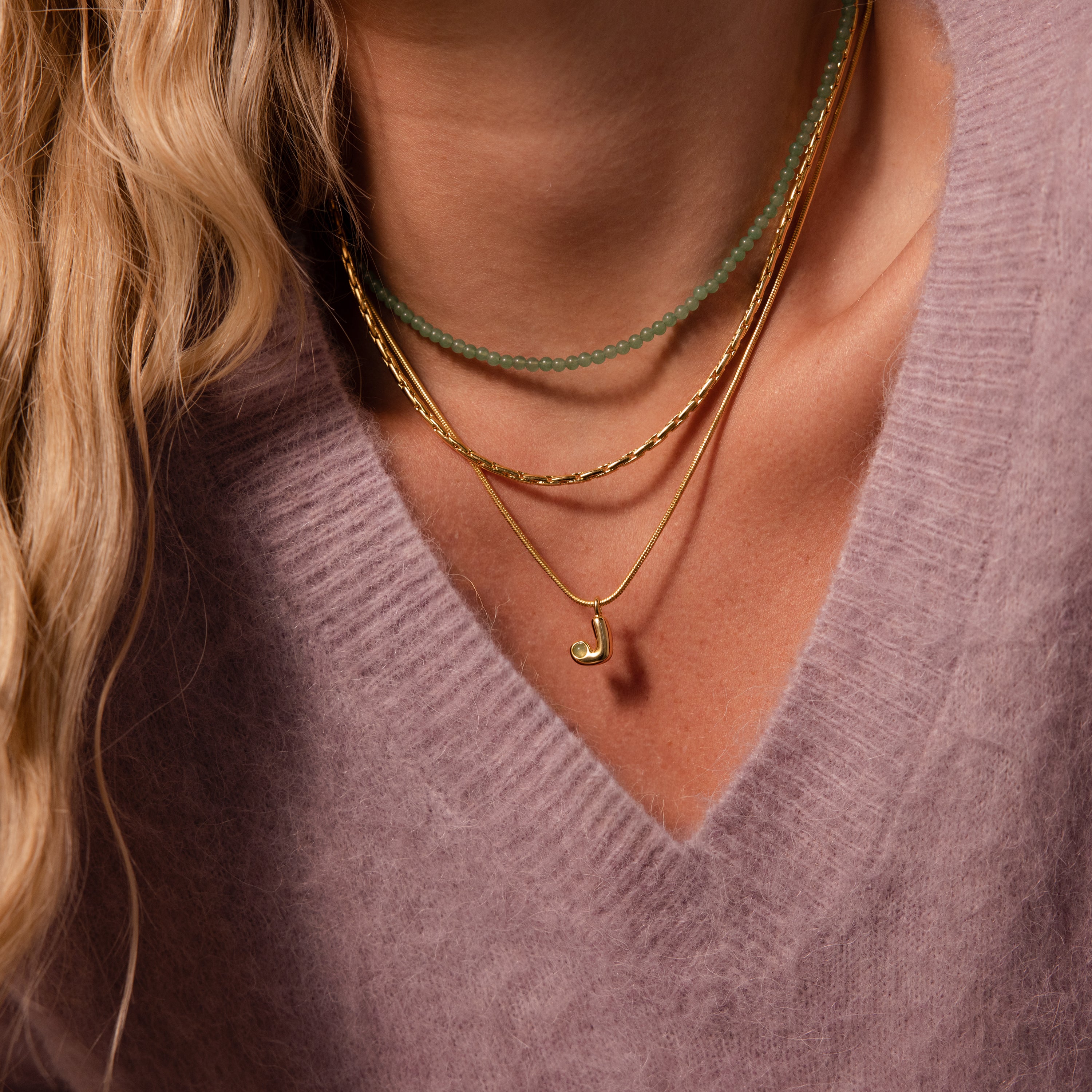 Close-up of a woman wearing a mauve sweater and layered gold and green bead necklaces, featuring the Gemstone Bubble Initial Necklace for a personalized touch.