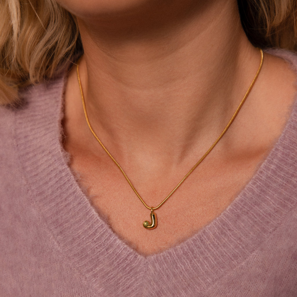 A woman wears the Gemstone Bubble Initial Necklace featuring a gold J pendant over a light purple fuzzy sweater.