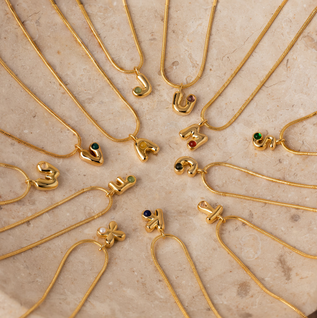 The Gemstone Bubble Initial Necklaces, adorned with vibrant gemstones, are displayed in a circle on beige marble, showcasing the elegance of personalized jewelry.
