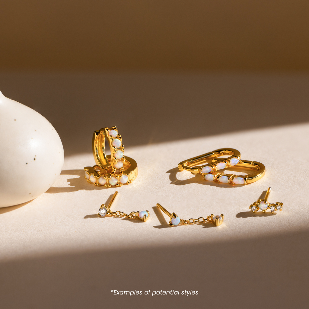 Four pairs of Mystery Jewelry gold and opal earrings are displayed on a light surface beside a white vase, each pair showcasing the whimsical charm and uniqueness of distinctive jewelry.