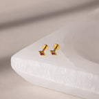 Gold geometric flat back ruby stud earrings on a white stand against a beige background