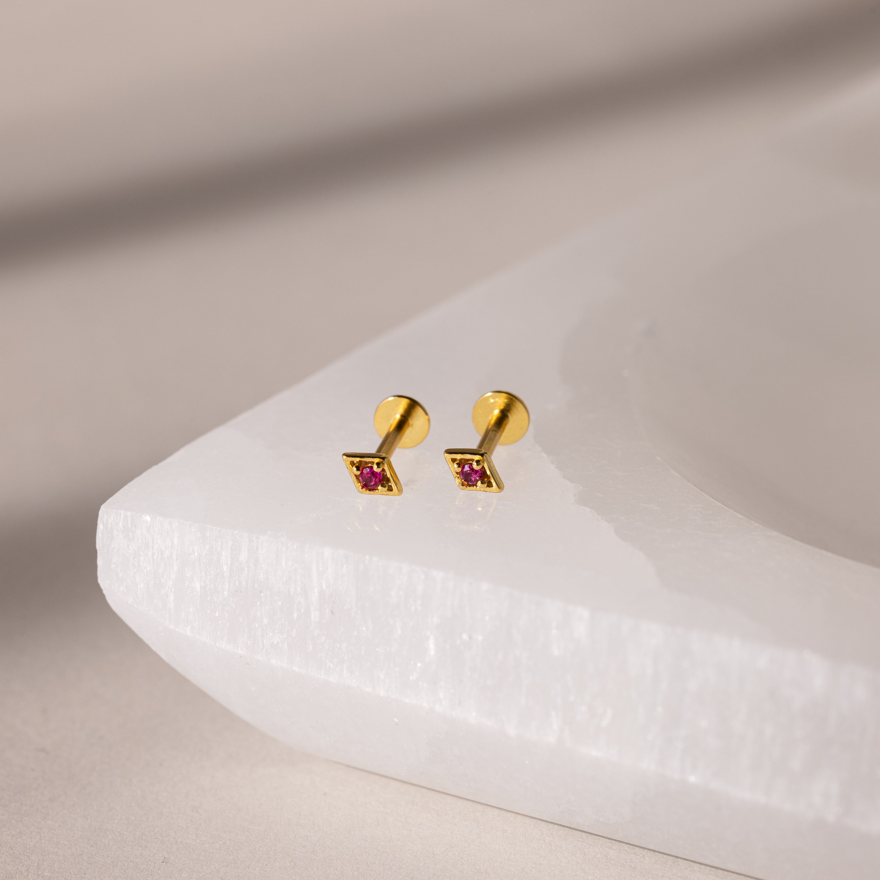 Gold geometric flat back ruby stud earrings on a white stand against a beige background