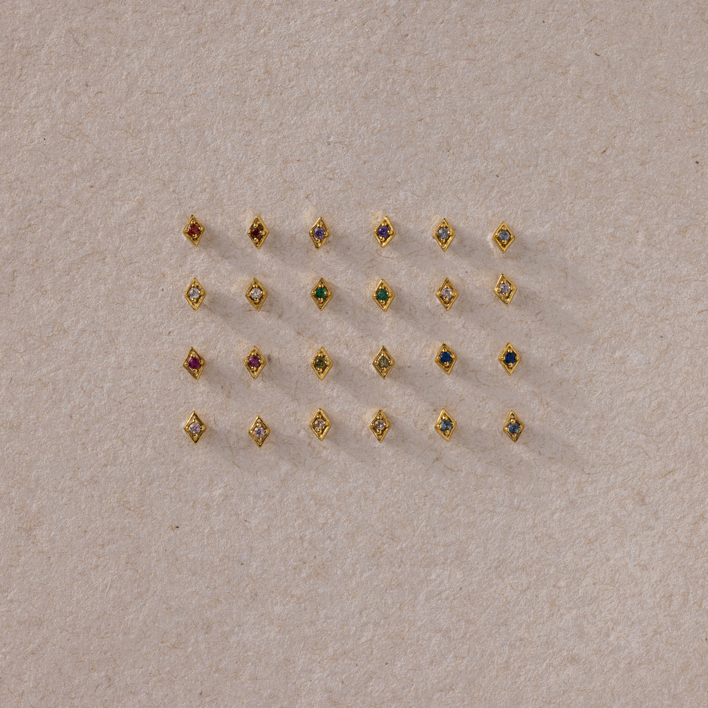 A flat lay of assorted gold geometric flat back studs arranged in a grid, featuring a variety of colorful gemstones on a neutral textured background.