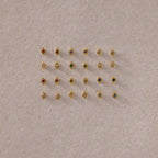 A flat lay of assorted gold geometric flat back studs arranged in a grid, featuring a variety of colorful gemstones on a neutral textured background.