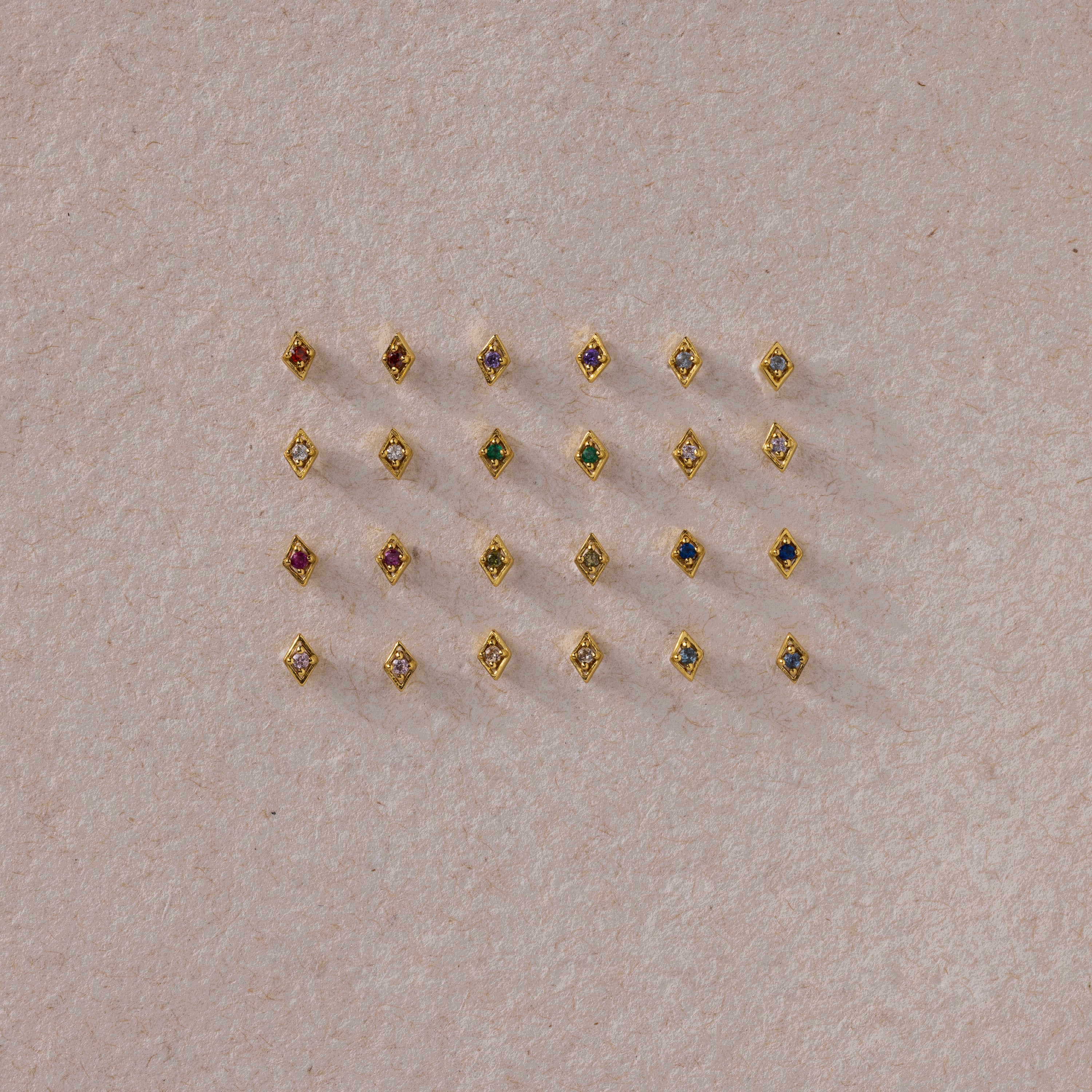 A flat lay of assorted gold geometric flat back studs arranged in a grid, featuring a variety of colorful gemstones on a neutral textured background.