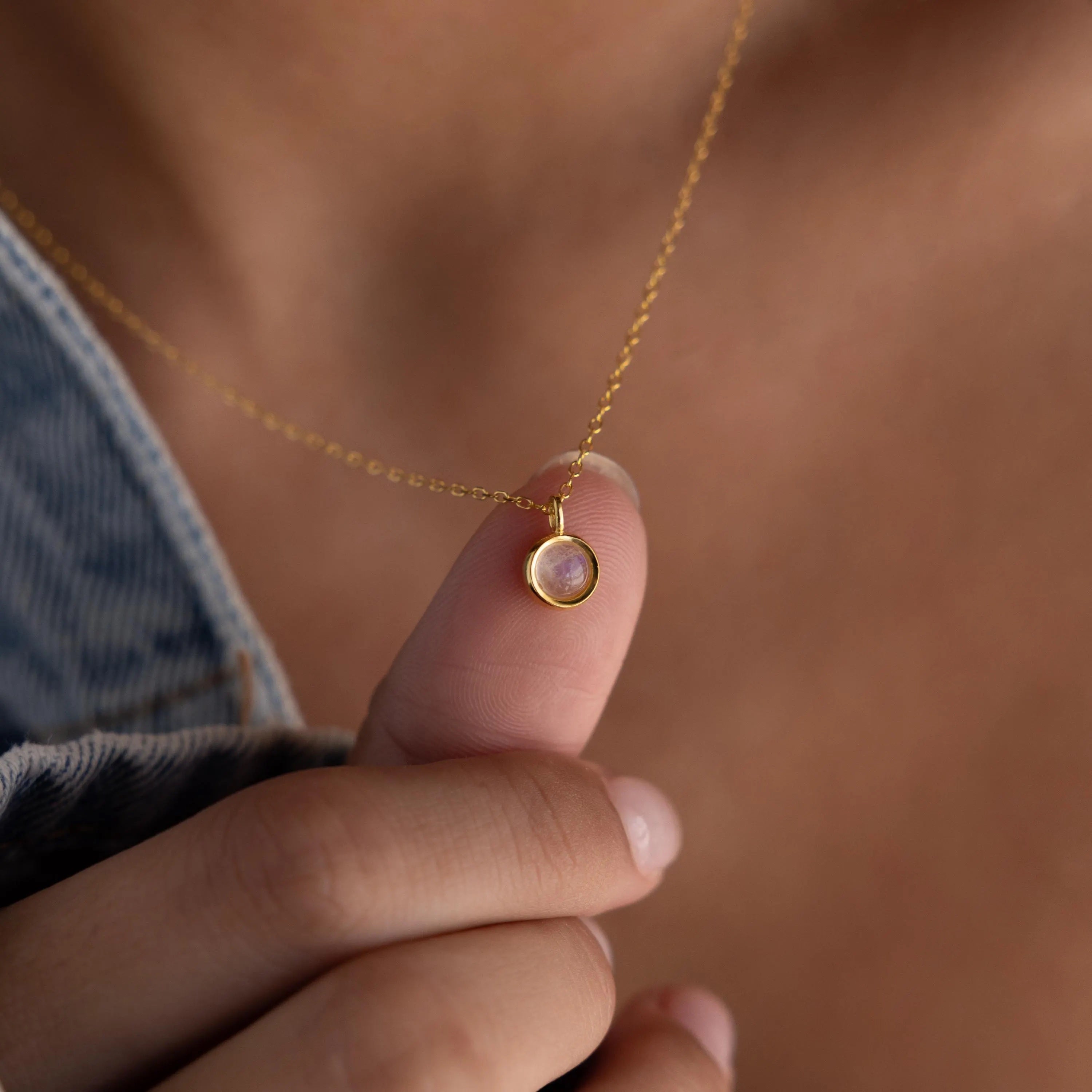 A hand holds the Moonstone Charm Necklace, its small round pendant featuring a shimmering moonstone that captures the ethereal beauty of this jewelry piece.