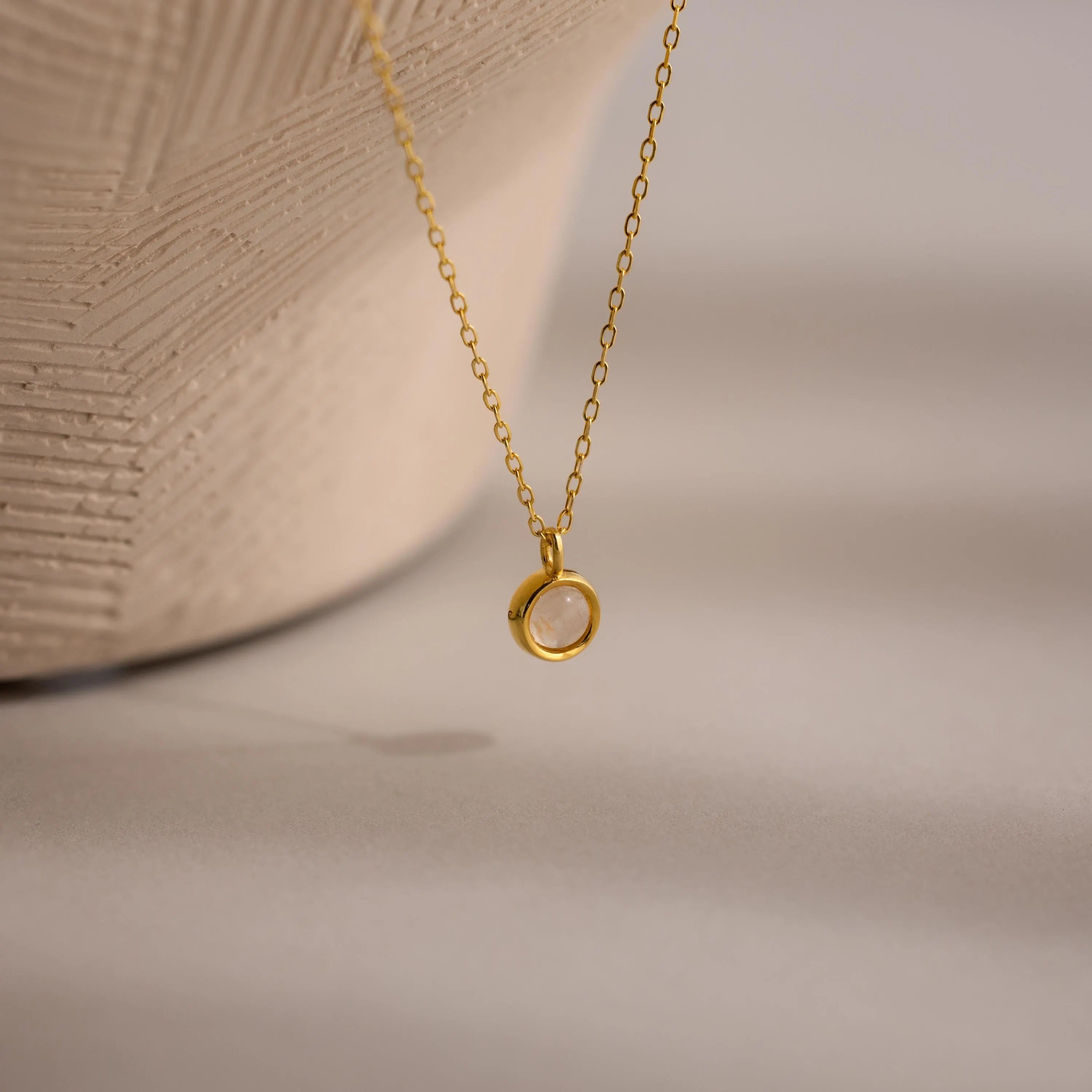 The Moonstone Charm Necklace features a gold chain with a small circular pendant, beautifully displayed on a neutral textured background—an elegant piece ideal for moonstone jewelry lovers.