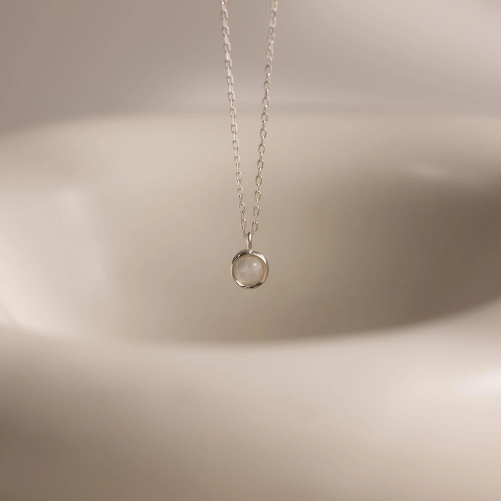 The Moonstone Charm Necklace features a delicate silver chain with a round, clear moonstone pendant—an elegant celestial accessory perfect for everyday wear.