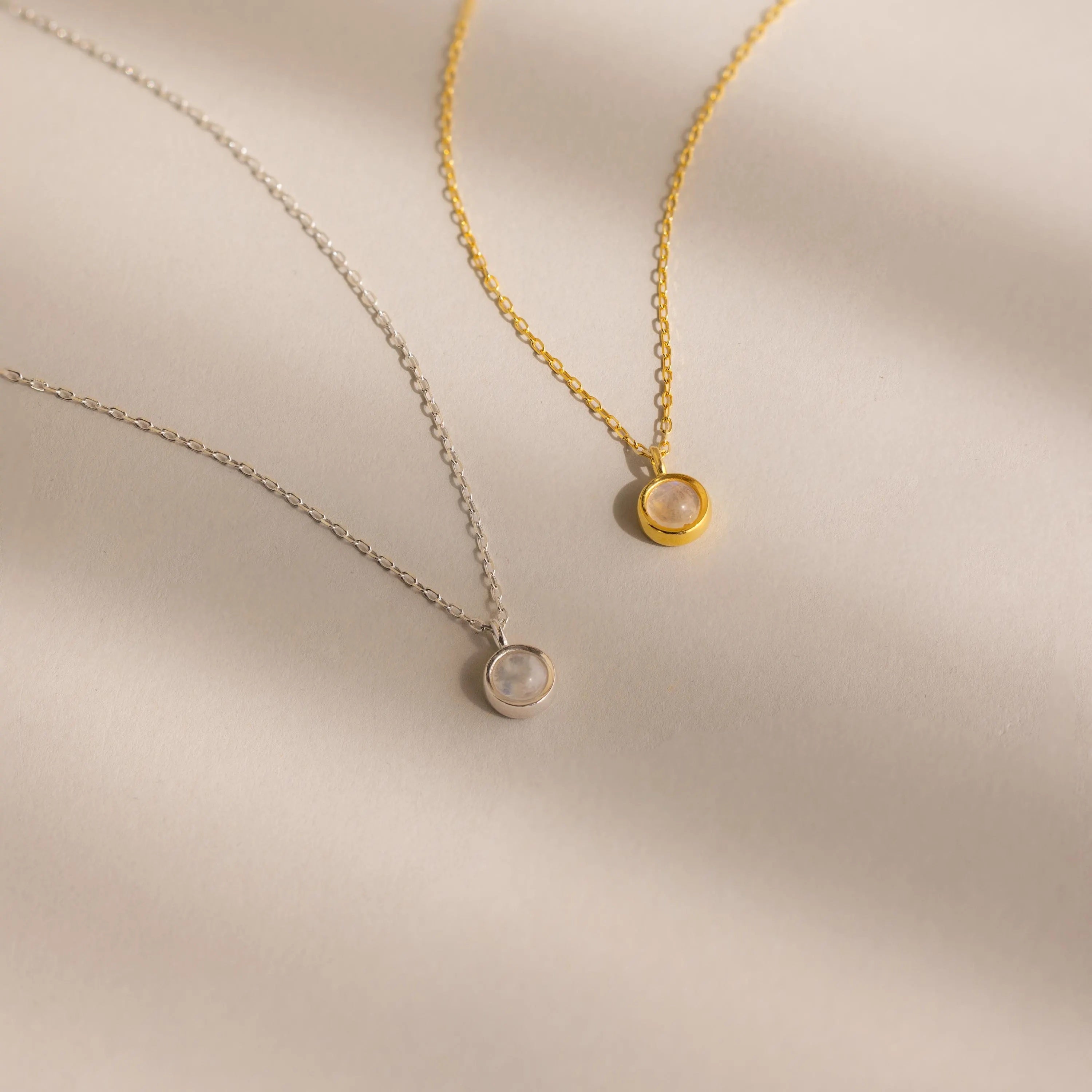 Layer these two delicate necklaces—one silver, one gold—each with a small round pendant on a light beige background. They’re perfect for stacking or pairing with your Moonstone Charm Necklace.