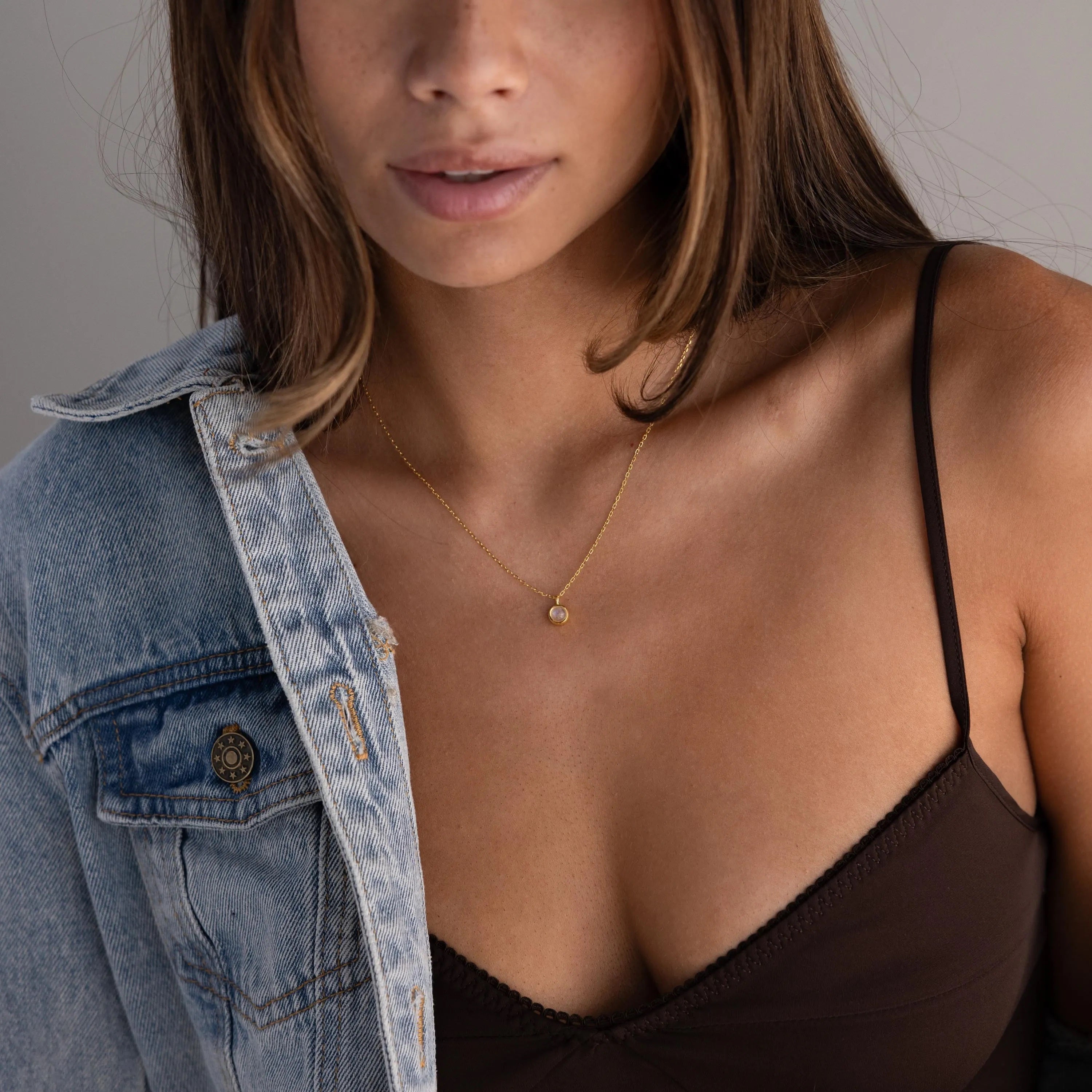 A woman in a black camisole and denim jacket wears the Moonstone Charm Necklace, featuring a delicate gold chain and a small celestial pendant.