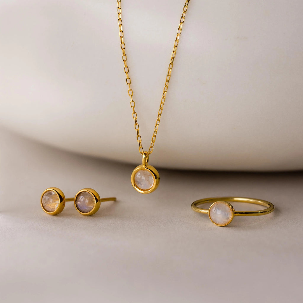The Moonstone Charm Necklace, paired with a matching ring and stud earrings, is displayed on a light surface—ideal for anyone who loves elegant gold moonstone jewelry.