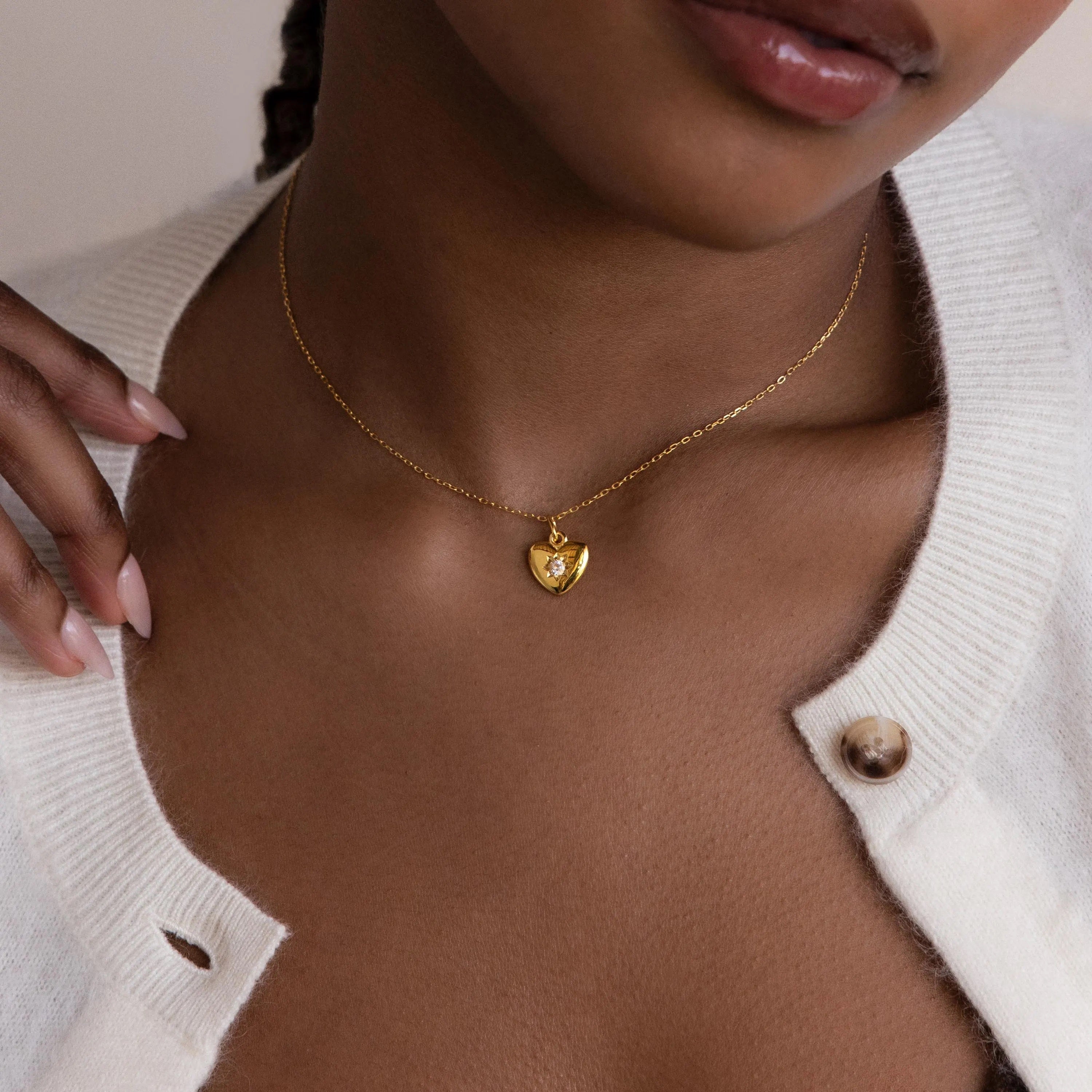 A woman wearing the North Star Birthstone Heart Necklace—a personalized gold heart-shaped pendant—and a white cardigan with an undone button.