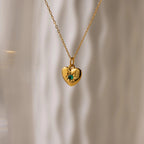 The North Star Birthstone Heart Necklace features a gold heart-shaped design with a green gemstone, shown against a soft, wavy light background.