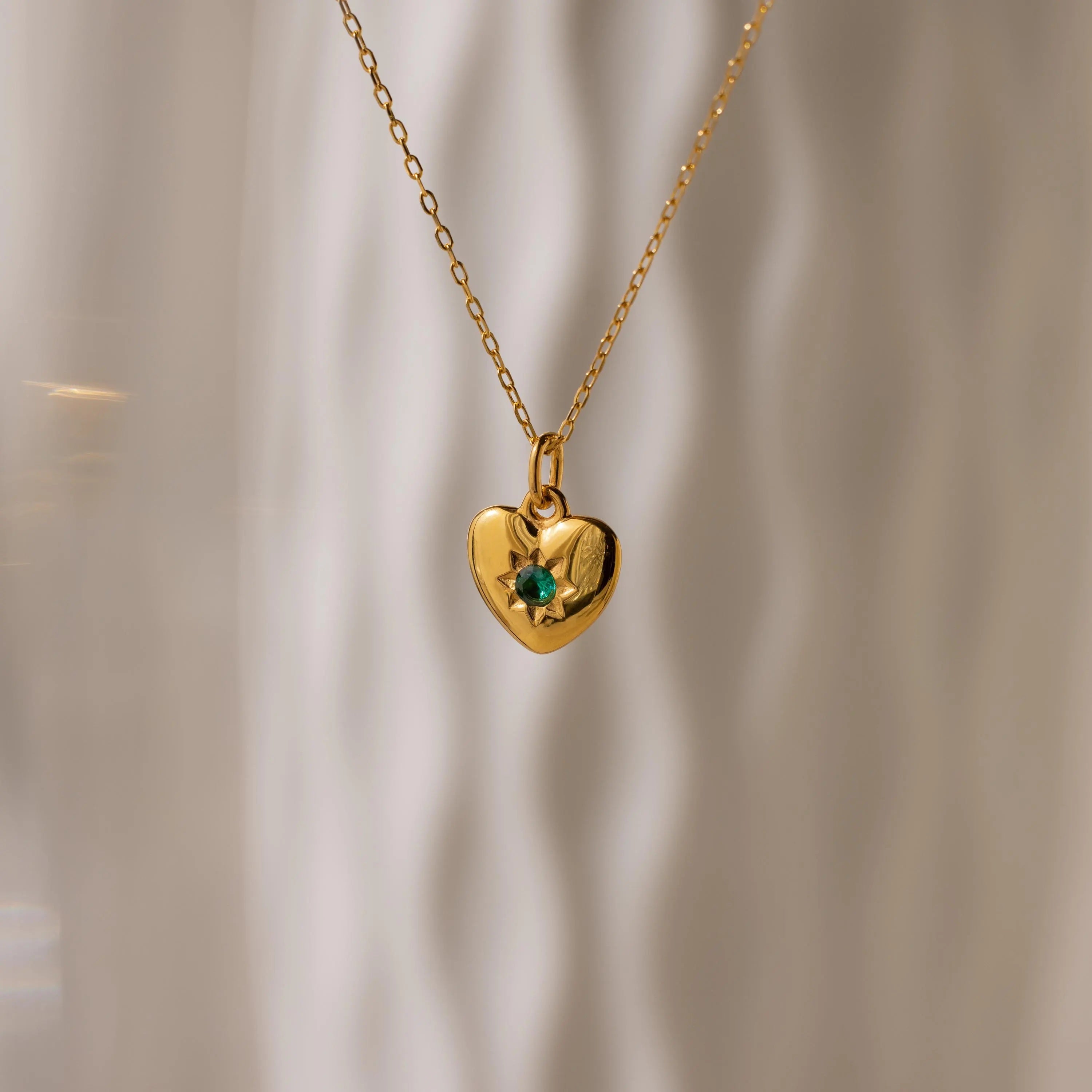 The North Star Birthstone Heart Necklace features a gold heart-shaped design with a green gemstone, shown against a soft, wavy light background.