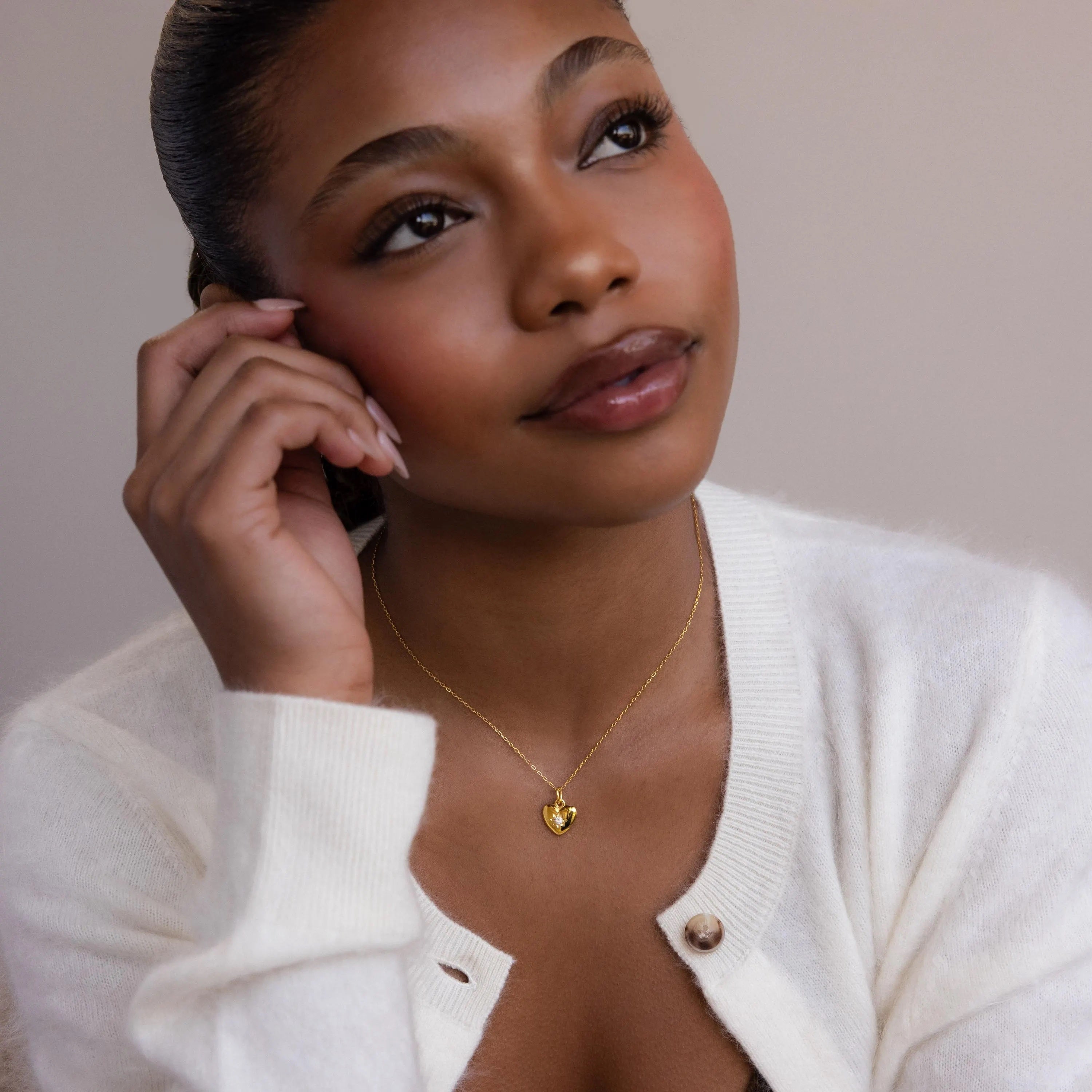 Woman in a white sweater wears the North Star Birthstone Heart Necklace, gazing up thoughtfully with her hand near her face.