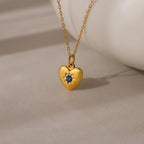 The North Star Birthstone Heart Necklace features a gold heart-shaped pendant with a blue gemstone on a delicate gold chain, displayed on a neutral surface.