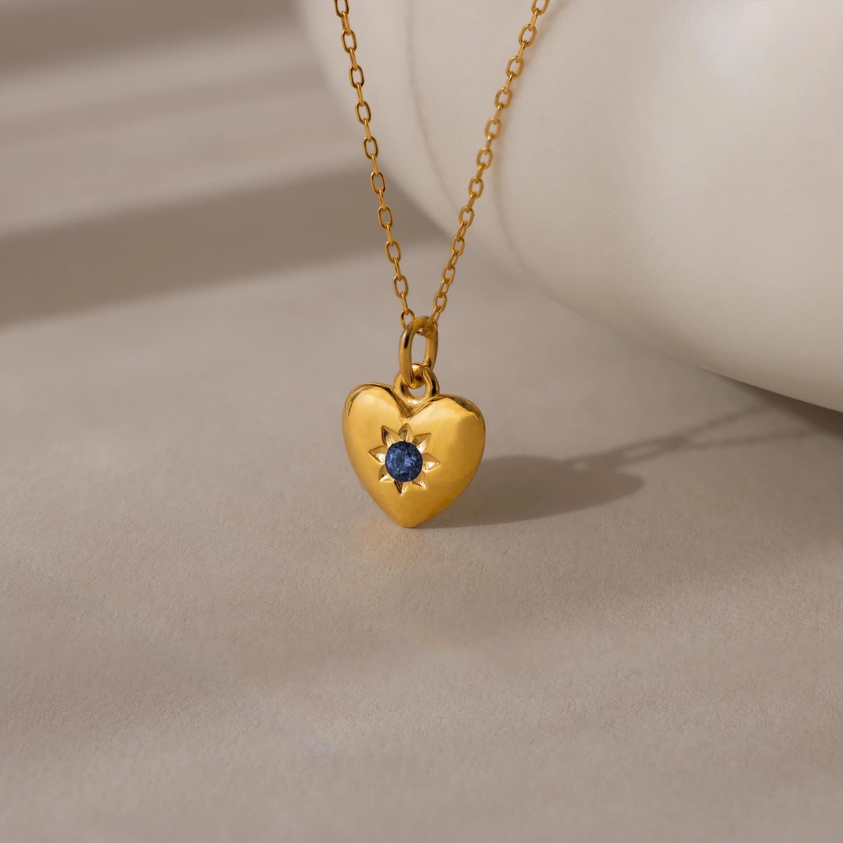 The North Star Birthstone Heart Necklace features a gold heart-shaped pendant with a blue gemstone on a delicate gold chain, displayed on a neutral surface.