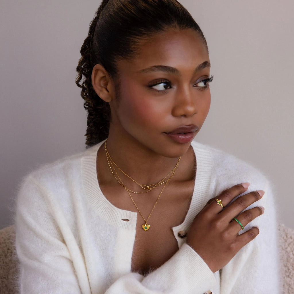 A woman in a white sweater wears gold rings and the North Star Birthstone Heart Necklace, looking to the side against a neutral background.