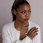 A woman in a white sweater wears gold rings and the North Star Birthstone Heart Necklace, looking to the side against a neutral background.