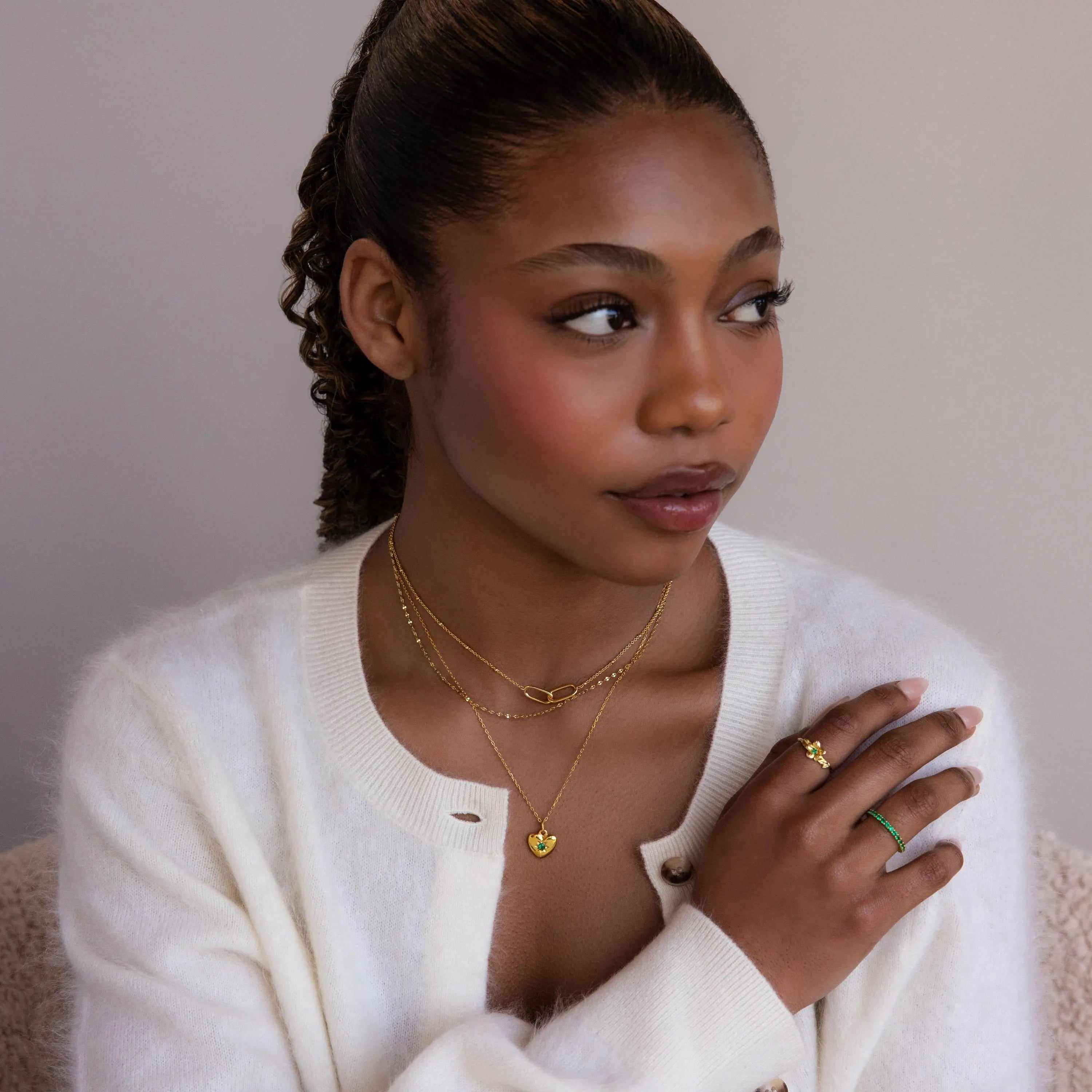 A woman in a white sweater wears gold rings and the North Star Birthstone Heart Necklace, looking to the side against a neutral background.