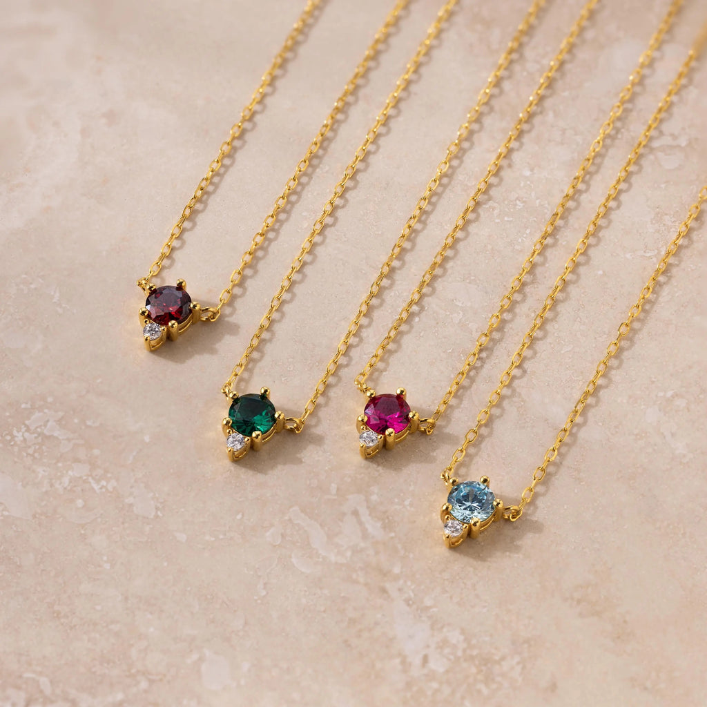 The Dainty Birthstone Drop Necklace features four gold chains with uniquely colored gemstone pendants, ideal for layering or as personalized birthstone jewelry, displayed on a light beige surface.
