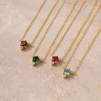 The Dainty Birthstone Drop Necklace features four gold chains with uniquely colored gemstone pendants, ideal for layering or as personalized birthstone jewelry, displayed on a light beige surface.