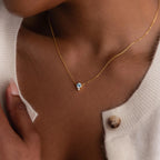 The Dainty Birthstone Drop Necklace with a blue pendant graces a woman's neck, styled with a cream cardigan and button.