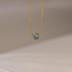 The Dainty Birthstone Drop Necklace features a gold chain with a blue gemstone pendant and a small clear stone, elegantly displayed against a neutral background.