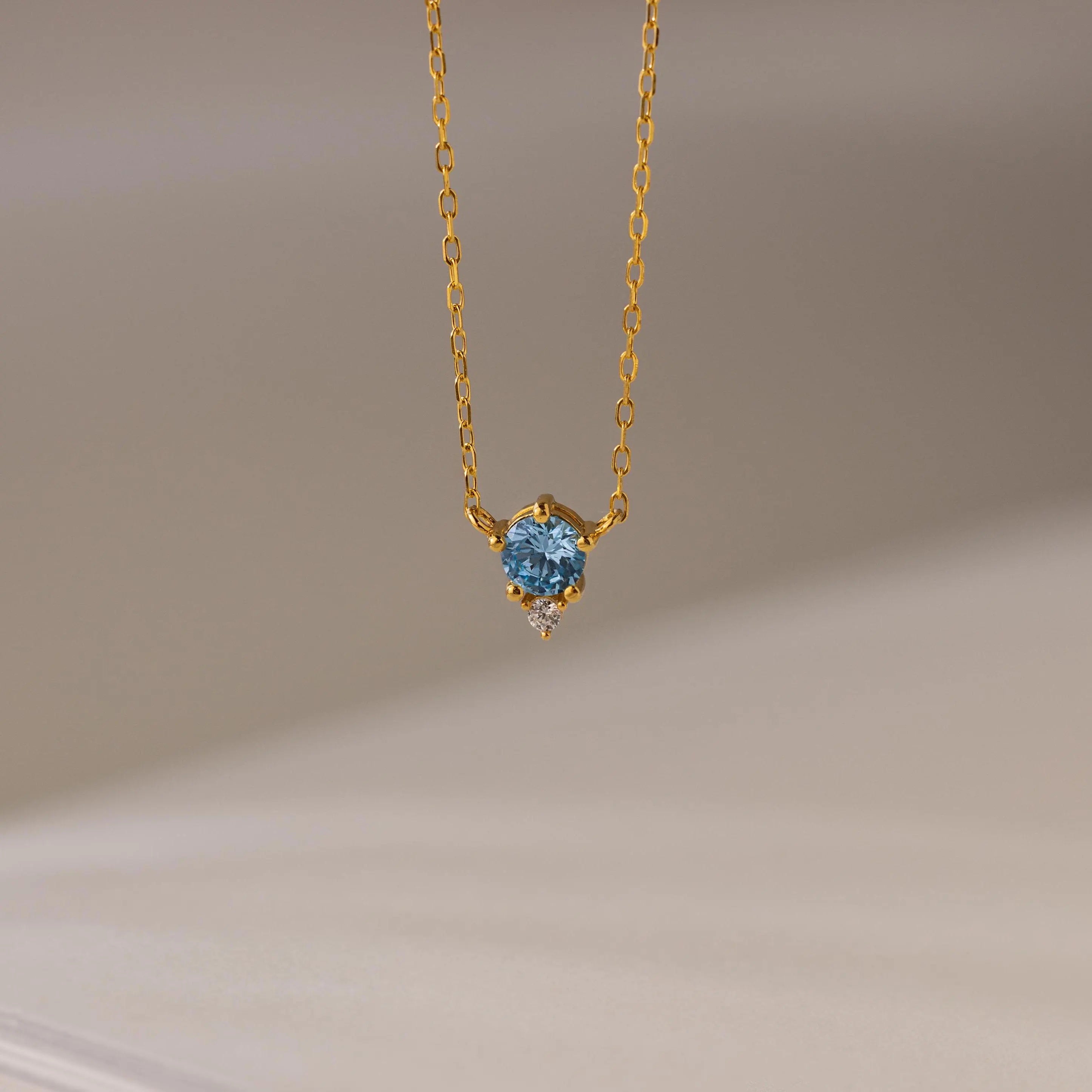 The Dainty Birthstone Drop Necklace features a gold chain with a blue gemstone pendant and a small clear stone, elegantly displayed against a neutral background.