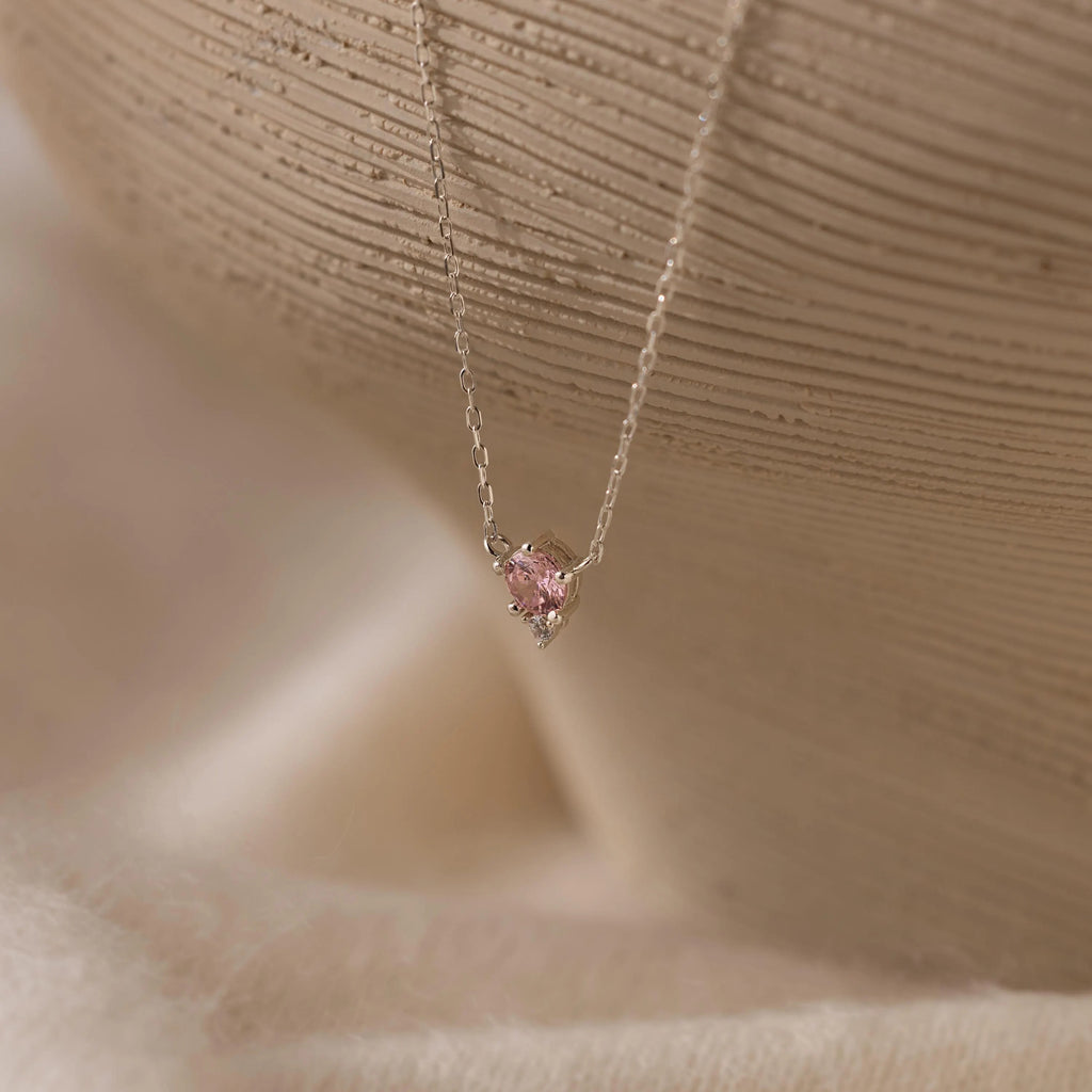 The Dainty Birthstone Drop Necklace showcases a small pink gemstone pendant set in delicate silver, presented against a soft, neutral background.