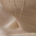The Dainty Birthstone Drop Necklace showcases a small pink gemstone pendant set in delicate silver, presented against a soft, neutral background.