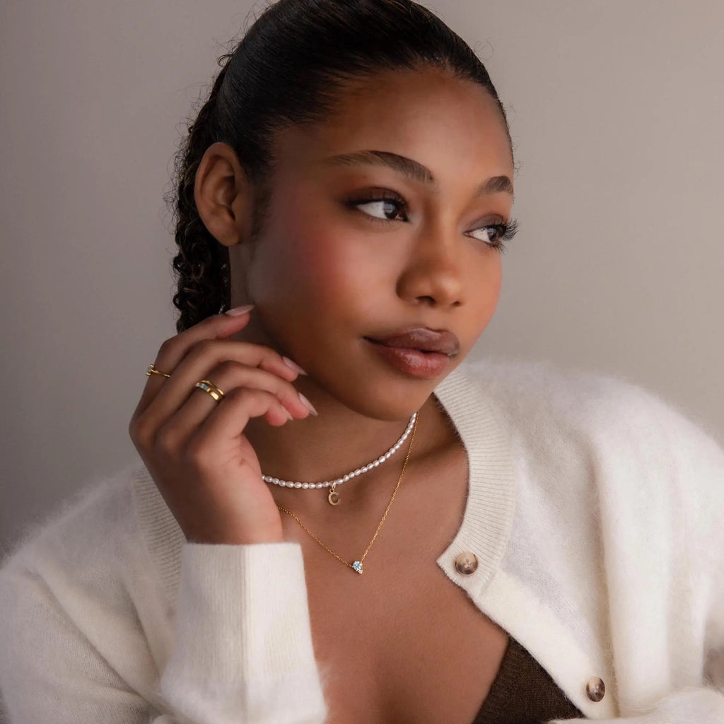 A woman with glowing skin wears a cream cardigan, gold jewelry, and the Dainty Birthstone Drop Necklace, her thoughtful expression gently turned to the side.