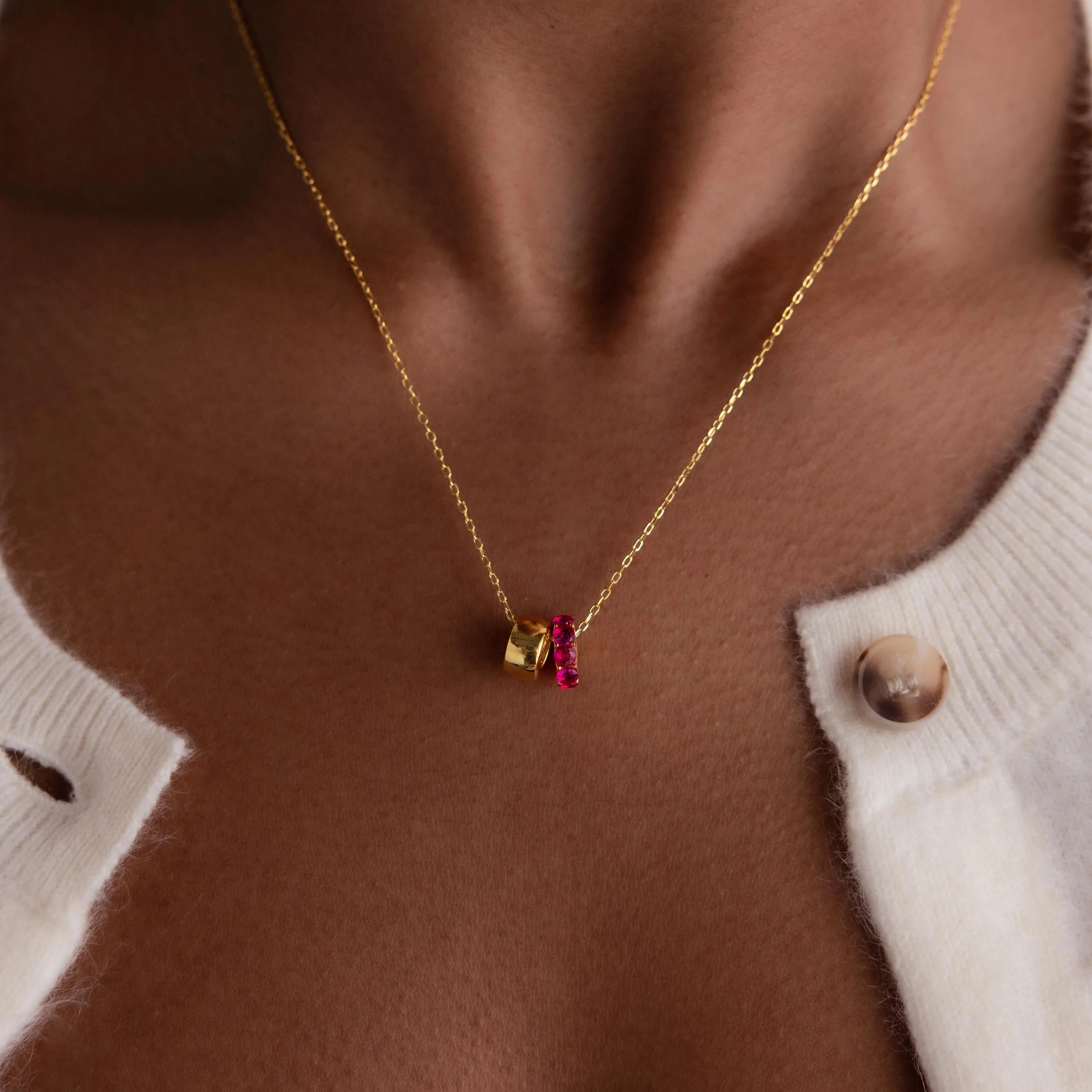 Close-up of the Duo Circle Birthstone Necklace, a gold chain featuring red and gold charms, including a circle charm, styled with a white buttoned sweater.