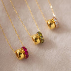Three Duo Circle Birthstone Necklaces with gold chains and red, green, and clear gemstones rest on beige fabric—ideal for those wanting personalized jewelry with elegant circle charms.