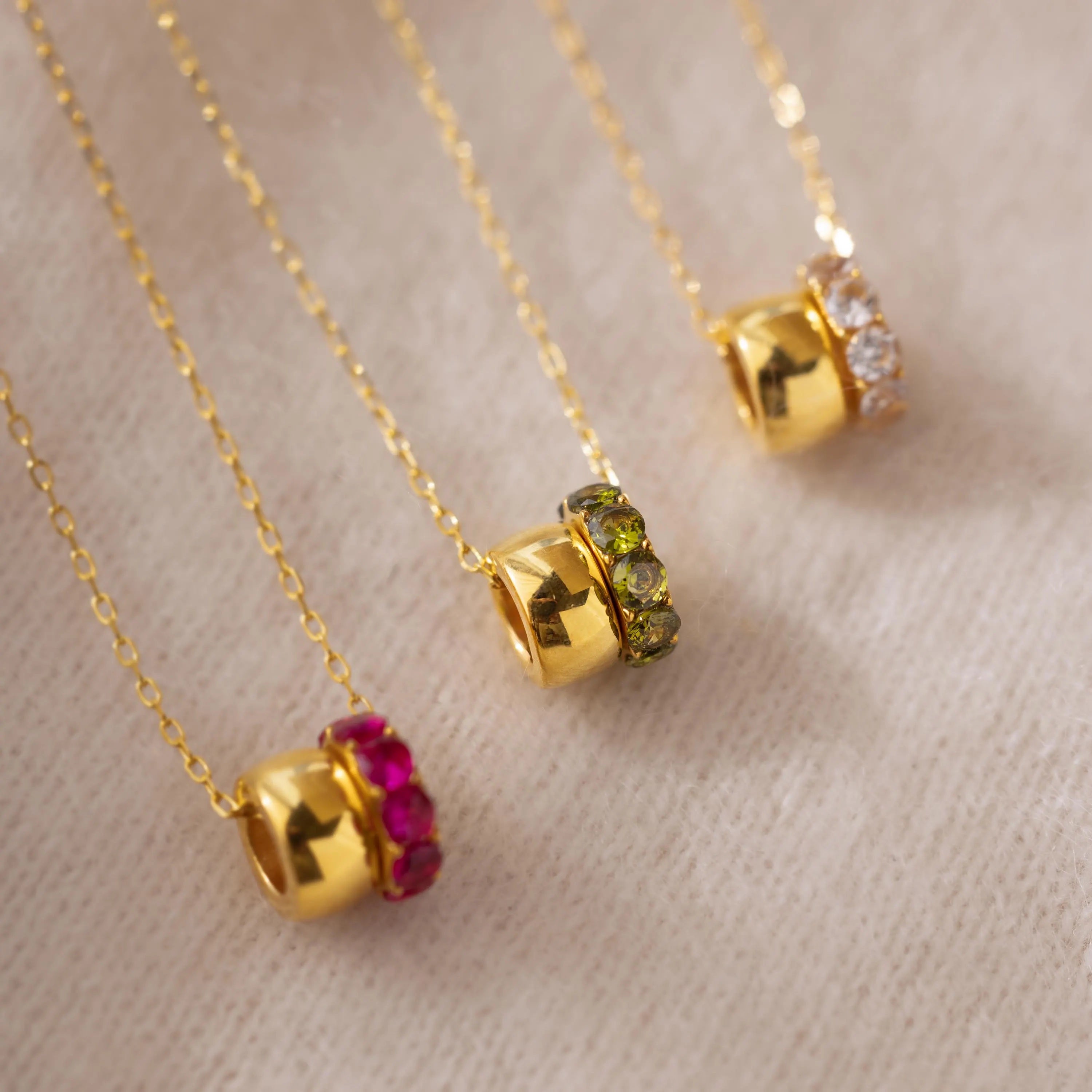 Three Duo Circle Birthstone Necklaces with gold chains and red, green, and clear gemstones rest on beige fabric—ideal for those wanting personalized jewelry with elegant circle charms.