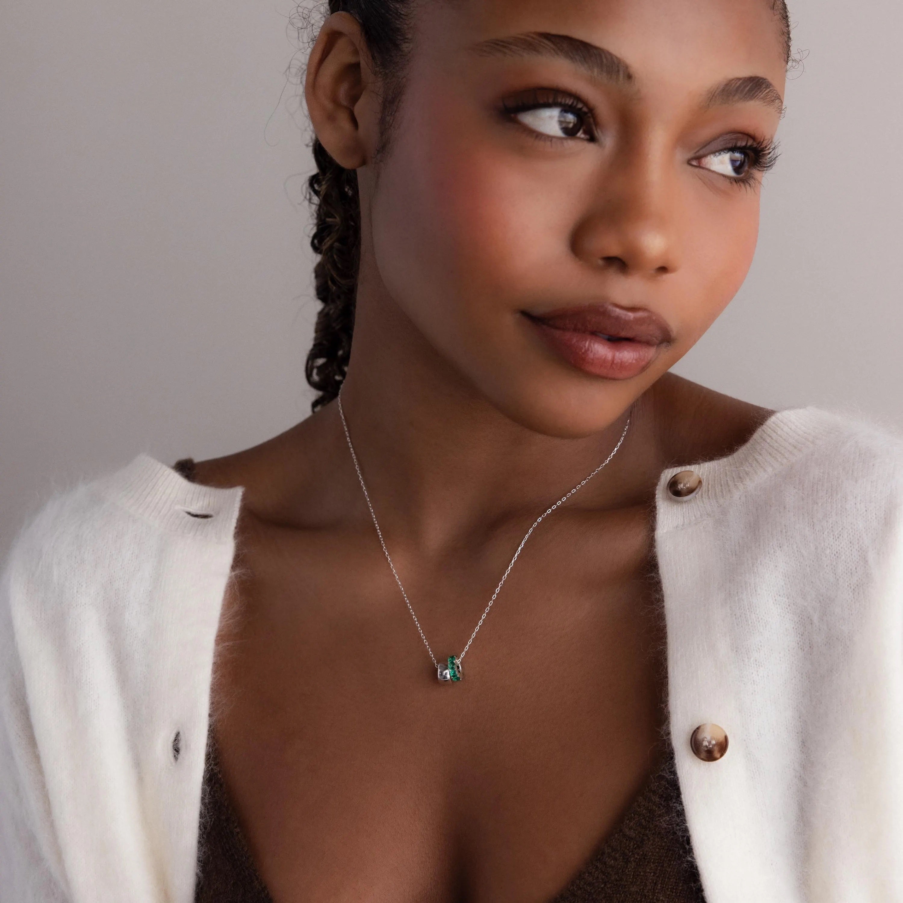 Woman in a white cardigan and brown top wears the Duo Circle Birthstone Necklace, featuring a green pendant, as she looks to the side.