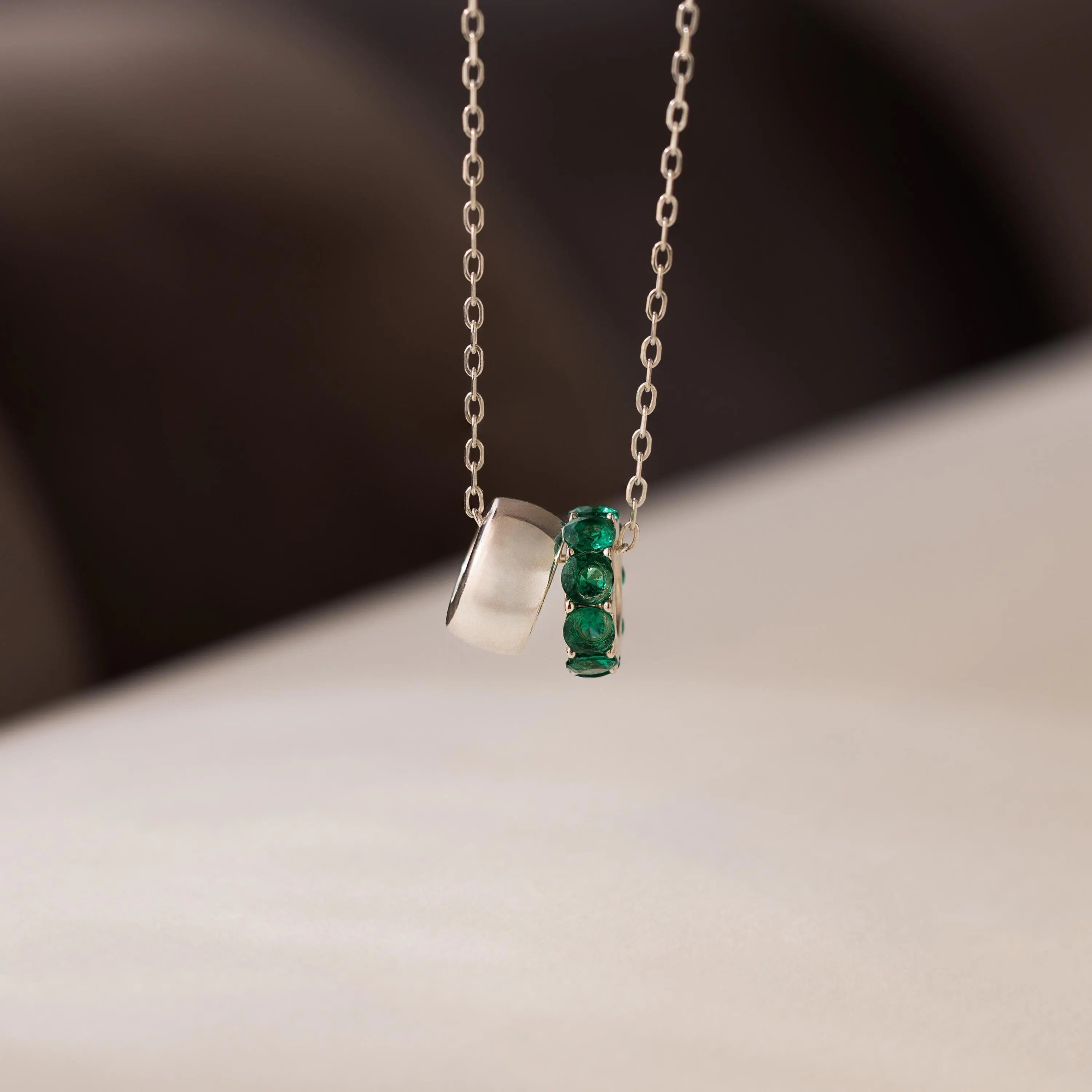 The Duo Circle Birthstone Necklace features two pendants: a matte silver circle and a green gemstone charm, set against a soft background.