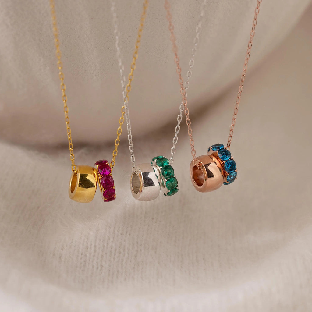 The Duo Circle Birthstone Necklace features gold, silver, and rose gold bands with colorful gemstones, beautifully displayed on soft fabric. Ideal for personalized jewelry lovers seeking elegance and meaning.