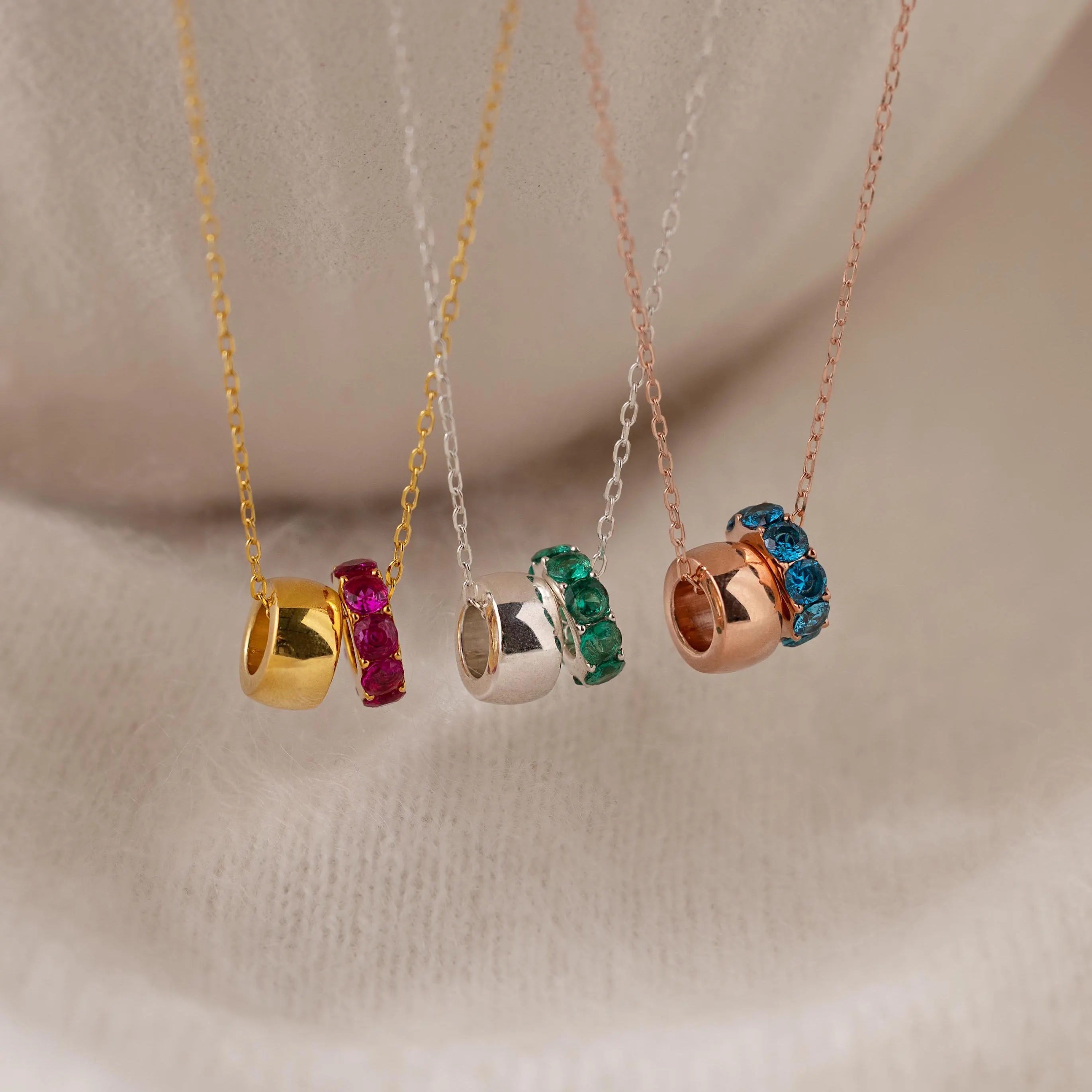 The Duo Circle Birthstone Necklace features gold, silver, and rose gold bands with colorful gemstones, beautifully displayed on soft fabric. Ideal for personalized jewelry lovers seeking elegance and meaning.