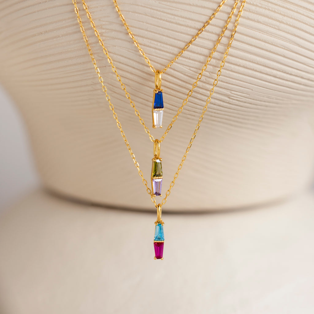 The Toi et Moi Tapered Birthstone Necklace, featuring a stunning baguette-cut pendant and colorful charms, is displayed on a cream textured stand alongside other gold necklaces.